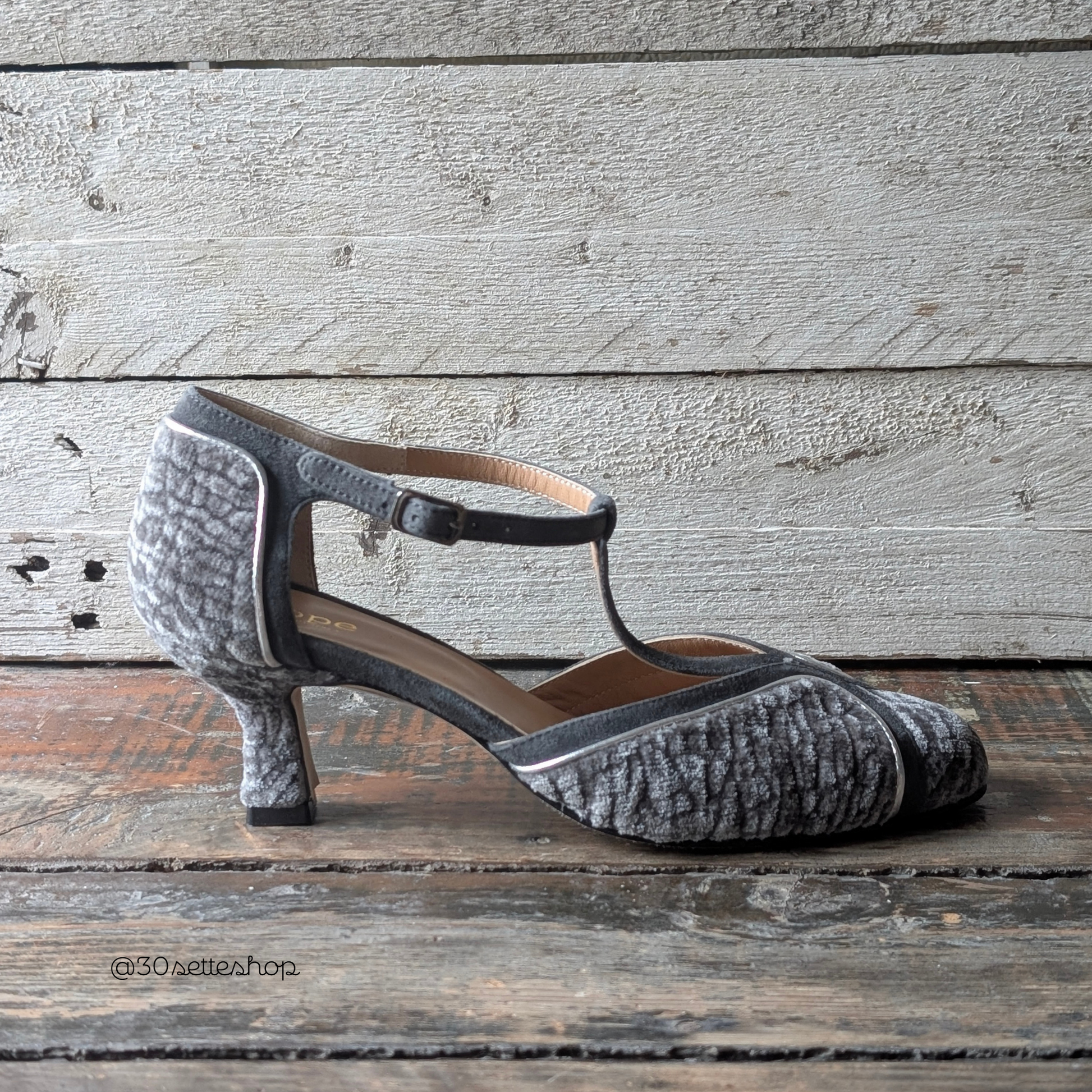 Bianca50 è una décolleté da donna con cinturino t-bar e tacco basso di 50 mm realizzata in un morbido velluto effetto astrakan color grigio perla abbinato al camoscio grigio del cinturino a T e al laminato argento del profilo sottile.