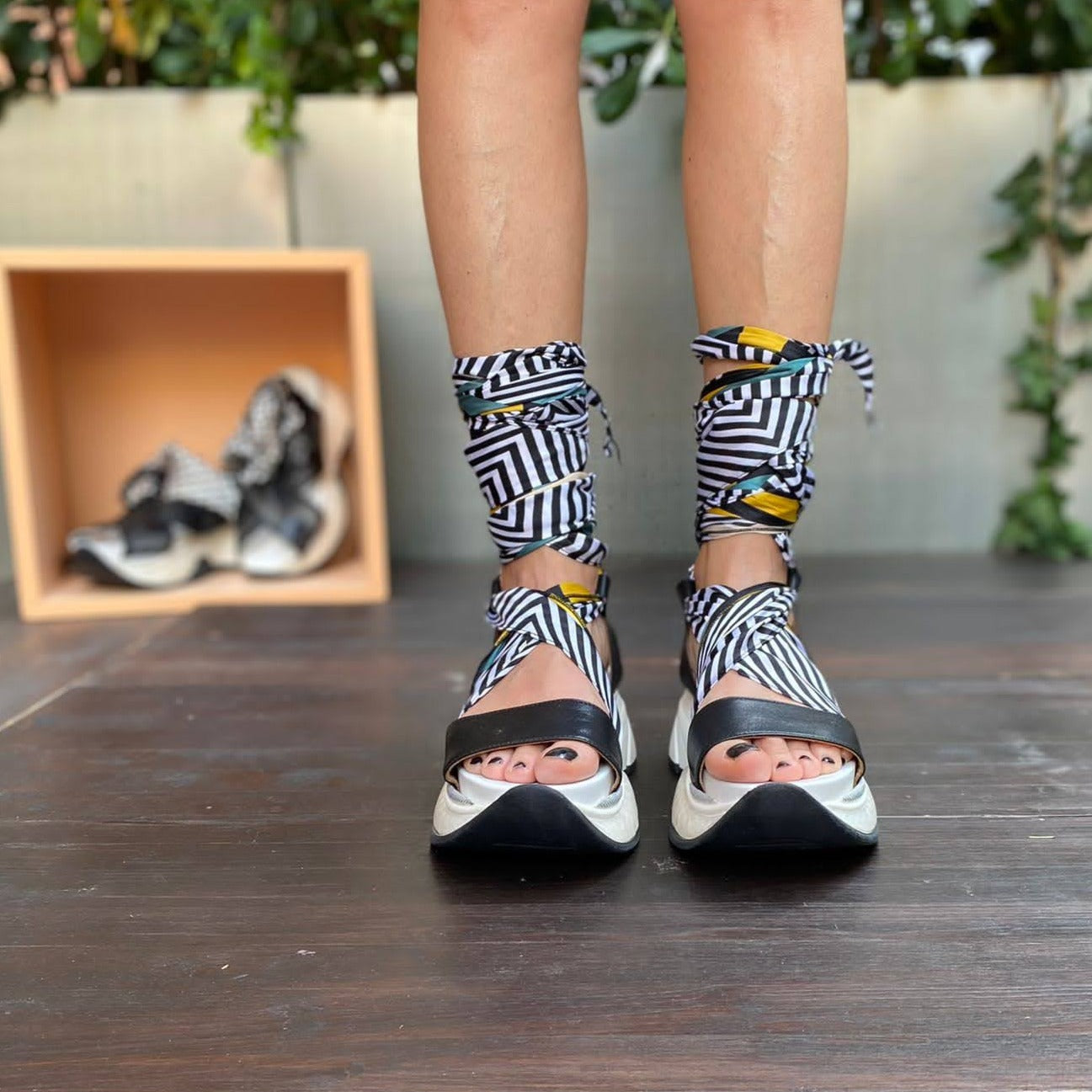Sneakers chunky o sandalo con zeppa in morbida pelle nera con foulard di seta montato tra suola e tomaia, che si intreccia sul piede e si avvolge alla caviglia. 